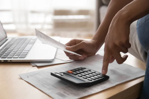 person using calculator and holding receipt paper in front of computer screen
